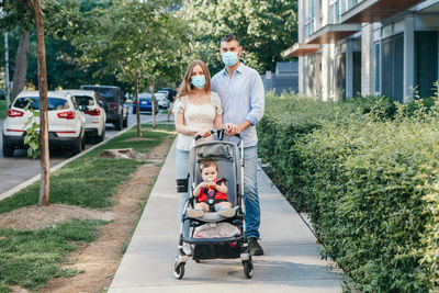 Caucasian mother and father in face masks walking with baby in stroller. 