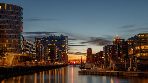 Harbour by illuminated buildings against sky at sunset