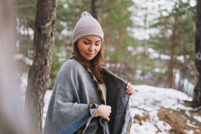 Portrait of young brunette beautiful woman in hat and grey poncho in the winter forest