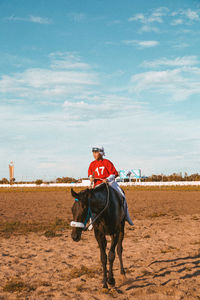 Rear view of young boy horse rider standing on field against clear sky