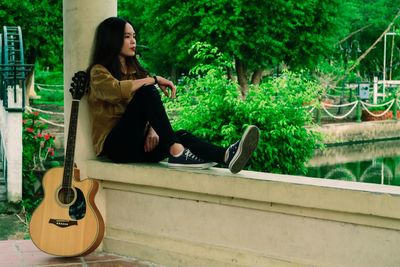 Full length of young woman sitting on retaining wall with guitar