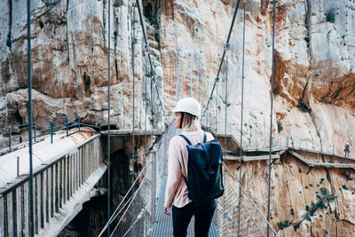 Rear view of young woman walking on rope bridge against rock