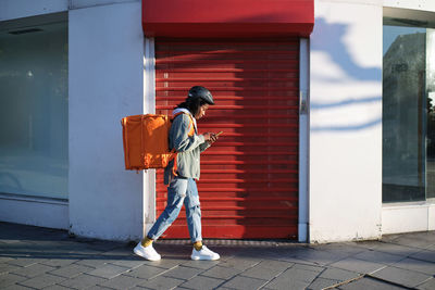 Full body side view of african american female with thermo backpack checking location on cellphone while walking along building in city