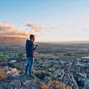 Young man standing on rock formation overlooking city against sky during sunset