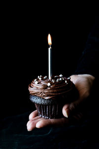 Hand holding a chocolate cupcake with a lit candle, against a black background.