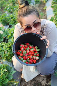 High angle portrait of woman wearing sunglasses showing strawberries in bucket at farm