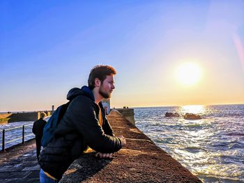 Young man leaning on retaining wall at beach against sky during sunset