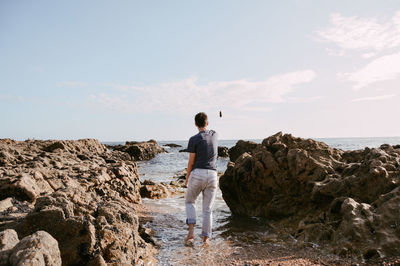 Rear view of young man standing at rocky shore against sky
