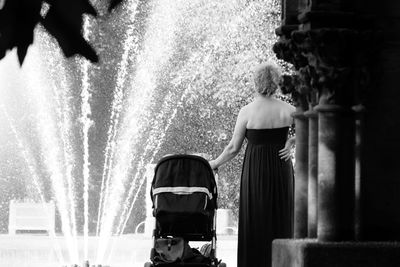 Rear view of woman standing with baby carriage against fountain