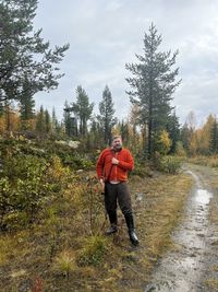 A bearded hunter in an orange sweater standing with his rifle by a forest road.