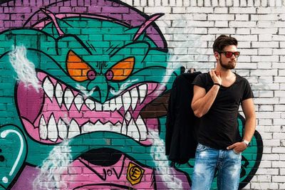 Young man wearing sunglasses standing against graffiti wall