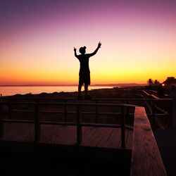 Silhouette man with arms outstretched standing on railing against sky during sunset