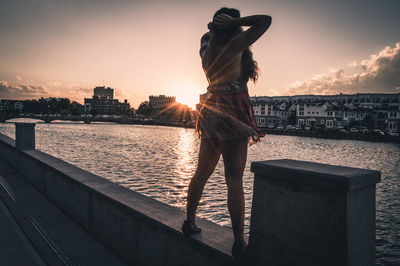 Rear view of young woman standing on retaining wall by river during sunset