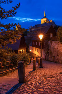 Illuminated street amidst buildings against sky at dusk