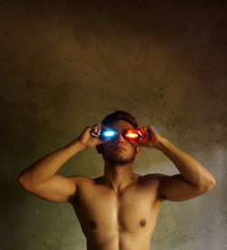 Shirtless muscular man holding illuminated neon lights on eyes by wall