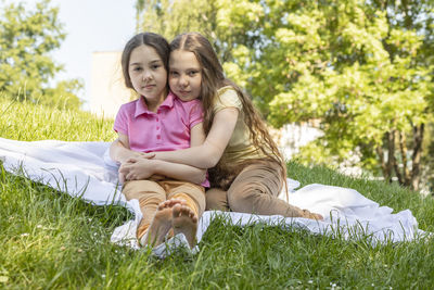 Two little girls, sisters with long hair sits hugging on grass in meadow at sunny day. caucasian