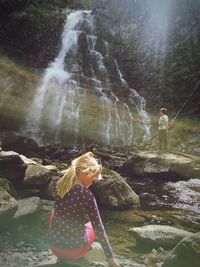 Full length of girl crouching on rock against waterfall