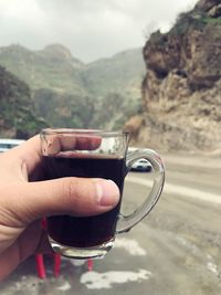 Close-up of hand holding black coffee in cup against mountains