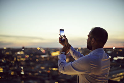 Mature businessman photographing illuminated city on smart phone while standing on office terrace during sunset