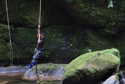 Full length of boy swinging on branch over lake against rocks