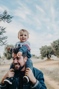 Side view of glad little boy riding on shoulders of smiling father during weekend in countryside