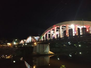 Illuminated bridge over river by buildings against sky at night