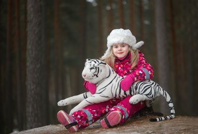 Portrait of girl in warm clothing holding toy tiger while sitting by trees in forest