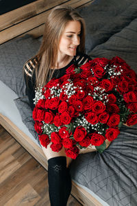 A beautiful girl is sitting on a bed in the bedroom with a huge bouquet of scarlet roses. 