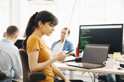 Female computer programmer using smart phone while sitting at desk in office