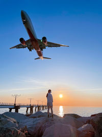 Rear view of man and airplane flying over sea against sky during sunset