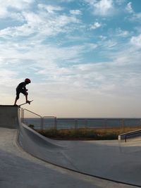 Young man performing stunt against cloudy sky at skateboard park