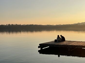 People sitting on pier over lake against sky during sunset