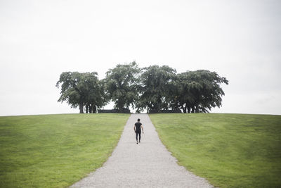 Mid distance view of young man walking on footpath amidst grassy land against trees and sky