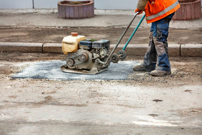 A worker with an old vibrating plate for road construction repairs potholes on an old road. 