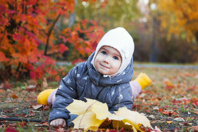 Cute smiling little girl is lying on a grass with yellow maple leaves on a walk in autumn park.