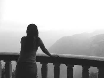 Rear view of young woman looking at mountains while standing by railing against sky during foggy weather