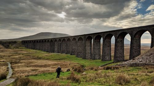 People on arch bridge on field against sky