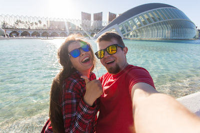 Portrait of young woman wearing sunglasses while standing against river