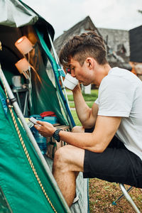 Young man drinking a coffee sitting at front of tent in the morning. teenager enjoying free time
