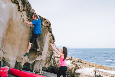 People climbing on rock at beach against sky