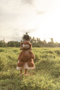 Girl carrying teddy bear on grassy field against sky