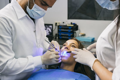 Male dentist in mask and gloves examining teeth of female patient with help of assistant at clinic