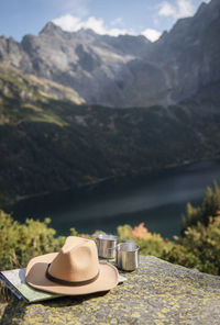Hot tea in metal tourist cup and hat on background of beautiful landscape of woods and mountains.