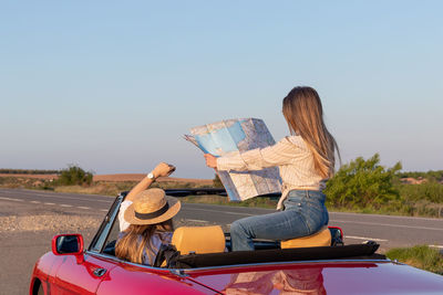 Women reading and holding a map sitting in red convertible car on remote road at sunset