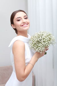 Side view of smiling woman with uptied hairstyle in wedding dress holding minimalist bouquet at home