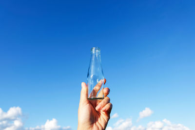 Cropped hand holding empty bottle against blue sky