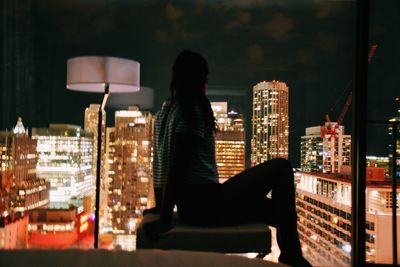 Woman on seat looking at illuminated buildings seen through window during night