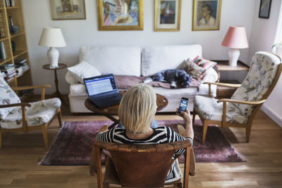 Rear view of senior woman using smart phone at home while dog relaxing on sofa