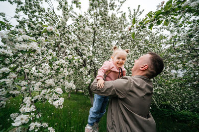 Dad holds a joyful baby daughter in his arms in a garden with a blooming apple tree