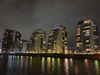 Illuminated buildings by river against sky at night